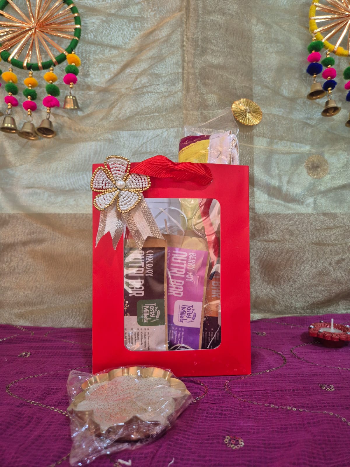 Red gift bag with three millet Nutri Bars, a bandhanwar, and traditional festive items.